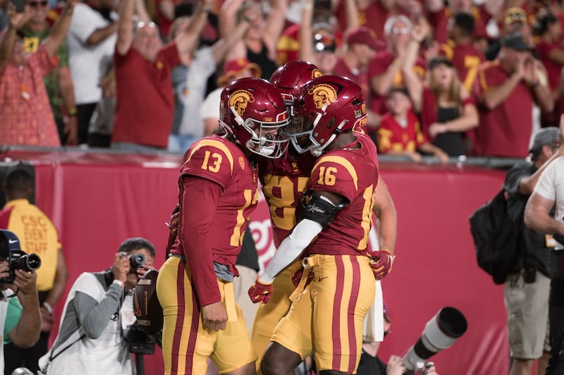 Caleb Williams (13) and USC teammates huddle in celebration. They wear cardinal football jerseys and helmets with gold accents and pants.