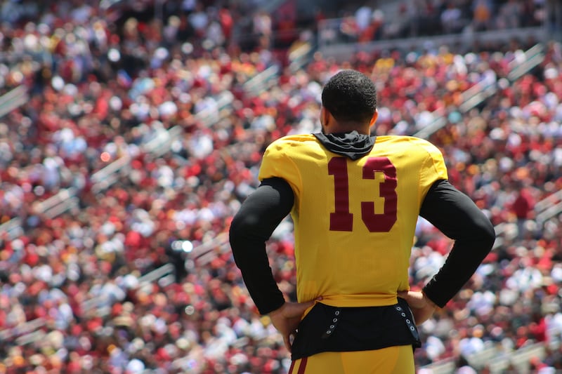 Caleb Williams (13) stands with hands on hips and back to the camera in the foreground; a large crowd of fans can be seen in the background. Williams wears a gold USC football jersey with cardinal accents on top of a long-sleeved black shirt.