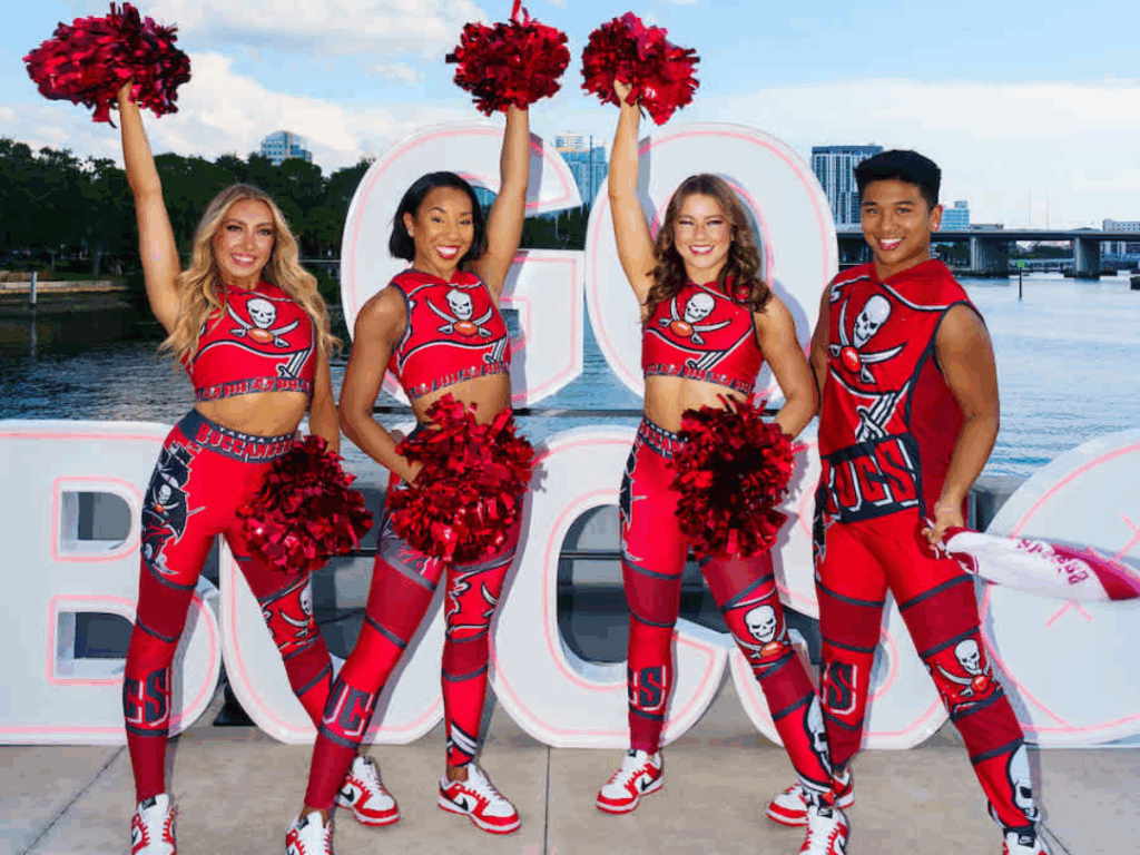 In front of a body of water and a sign that says "GO BUCKS," four cheerleaders pose for a photo. On the left are three women holding poms. On the right is a man swinging a rally towel.