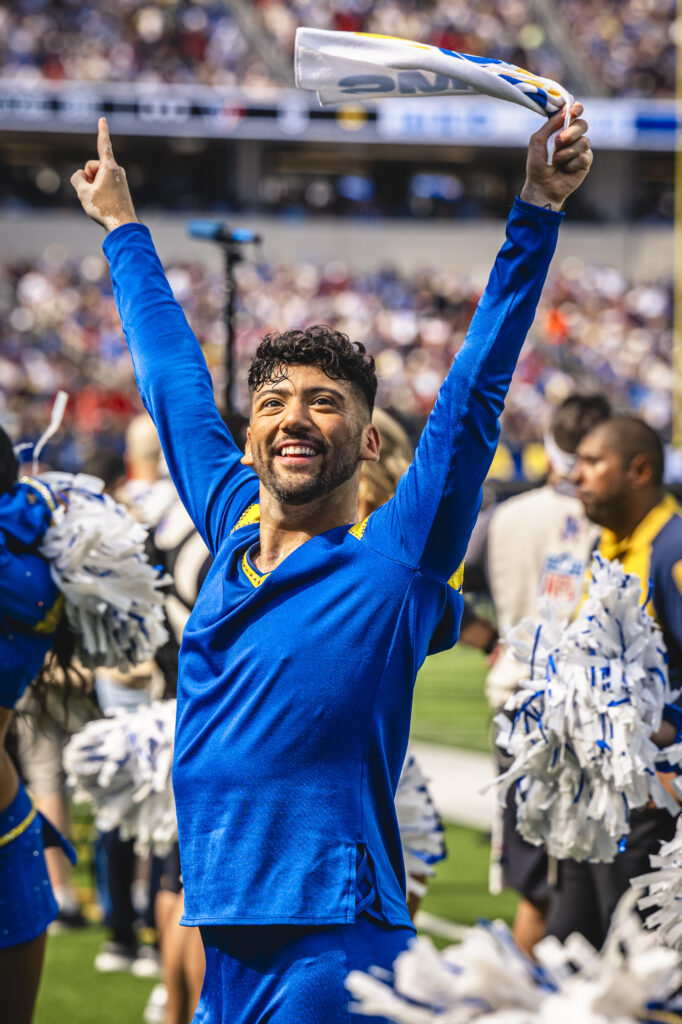 Jose Capetillo performs on the field as a Los Angeles Rams Cheerleader. He raises both arms up in a high "V," holding a rally towel in his left hand.