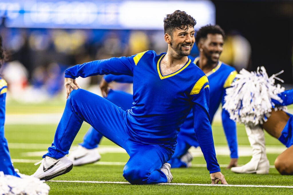 On a football field, the Los Angeles Rams Cheerleaders perform in blue uniforms. Jose Capetillo is shown in the middle. They sit on one leg, tucked under, with the other leg posed to the side on forced arch. They rest one arm on top of the extended leg with the other on the ground to support them.