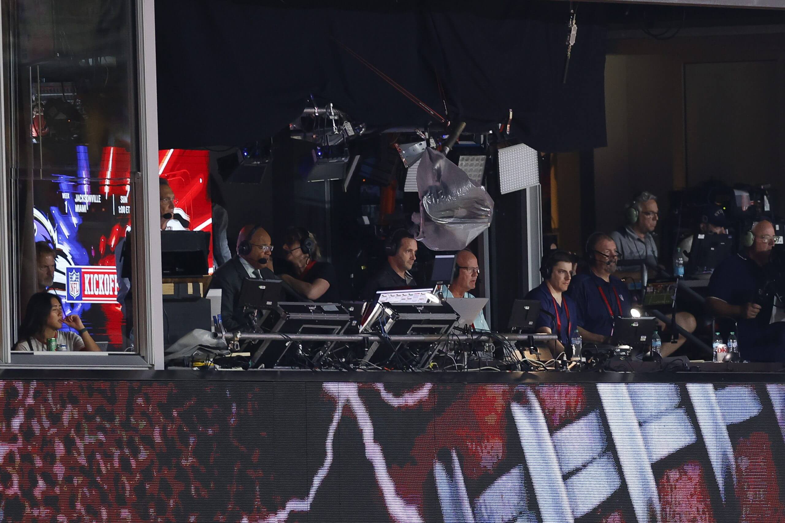 A view of the NBC broadcast booth from across the stadium, with at least a dozen people wearing headsets and looking at the field, with equipment — lights, cameras, monitors, microphones and much more — all around them.