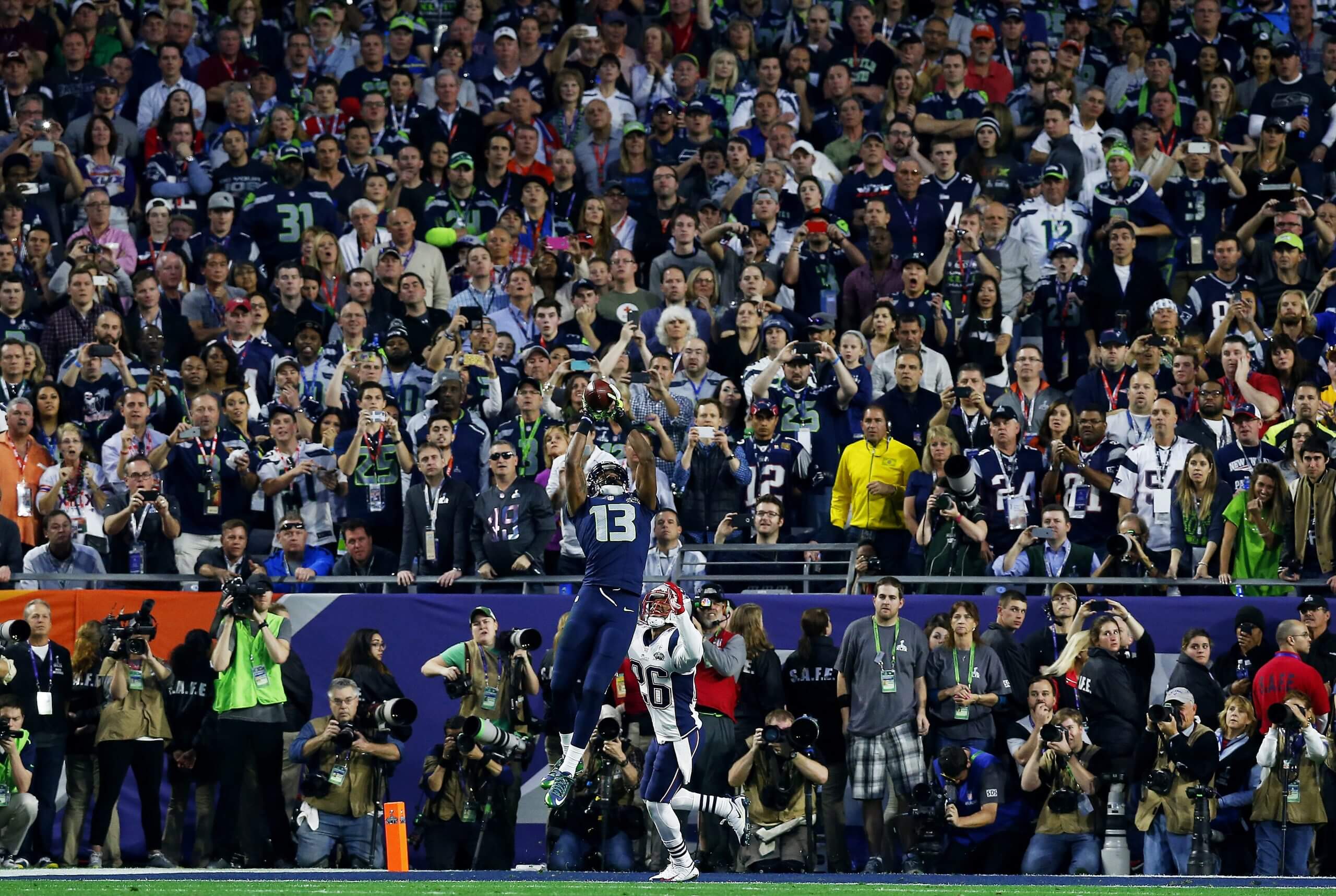 A wide shot showing Chris Matthews, in a navy No. 13 uniform, leaping high and snagging the ball with both hands up over his head, while Logan Ryan, in a white No. 26 uniform, looks up from below and dozens of photographers and fans look on in the background.