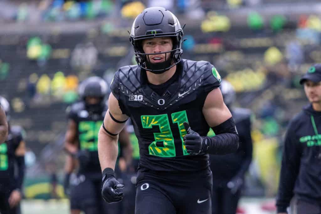 Oregon Ducks football player Dillon Thieneman wearing a black uniform with green numbers and a black helmet runs forward during warmups on a rainy day, with teammates blurred in the background.