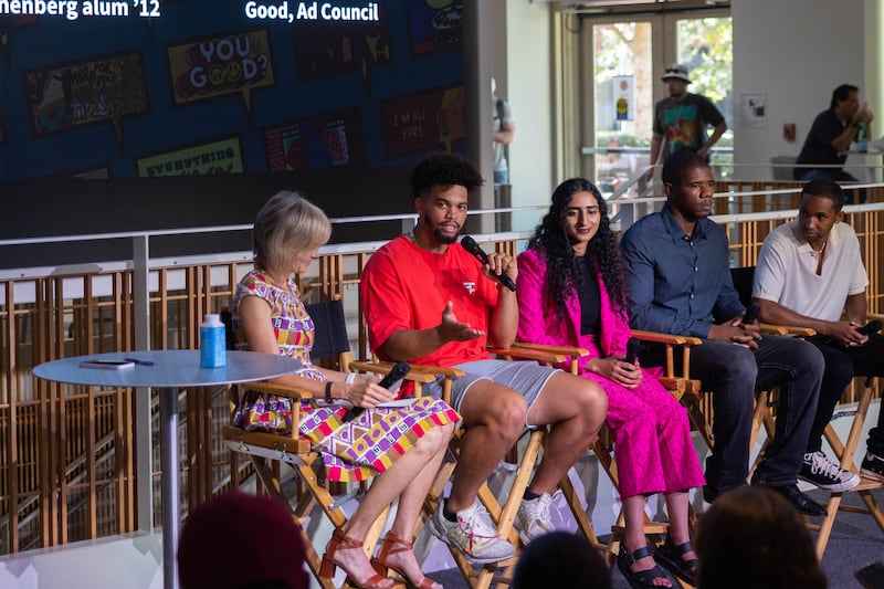 Caleb Williams sits in a director's chair amidst a panel of other contributors. He gestures to the crowd while holding the microphone and wears a bright orange shirt with gray shorts.