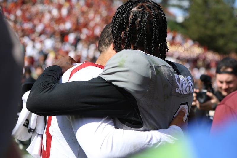 Caleb Williams and Shedeur Sanders hug after a USC/Colorado football game. Williams wears w hite football jersey with cardinal and gold accents, while Sanders wears a gray jersey with black and white accents.