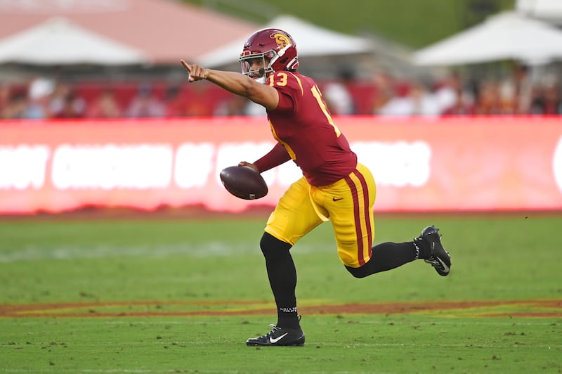 Caleb Williams (13) holds the football in his right hand and points forward with his left as he runs down the field. He wears a cardinal USC football jersey with gold accents and gold pants with black cleats.