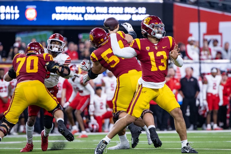 Caleb Williams (13) with arm outstretched to throw the football in his hand; other USC football players can be seen blocking Utah defenders in the background. They wear cardinal football jerseys and helmets with gold accents and pants.