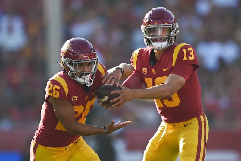 Williams (13) hands the football off to running back Travis Dye (26). Both wear cardinal USC football jerseys and helmets with gold accents and pants.