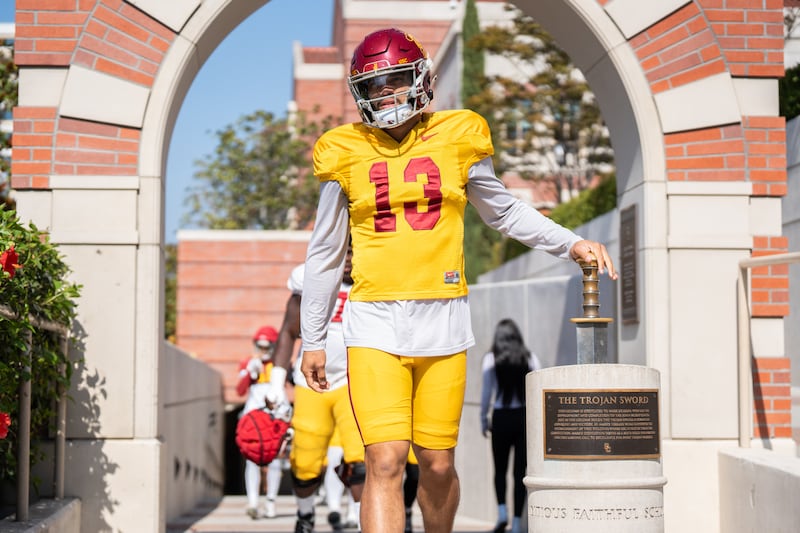 Williams (13) touches the Trojan Sword with his right hand as he emerges from the locker room before practice; he wears a gold USC football uniform with cardinal helmet.