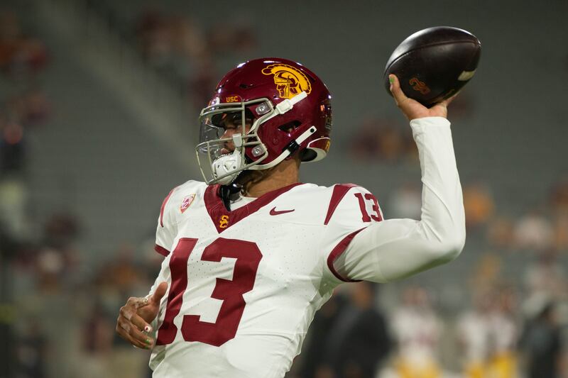 Caleb Williams (13) in profile with left arm extended back as if about to throw the football in his hand. He wears a white USC football jersey with cardinal accents and helmet.