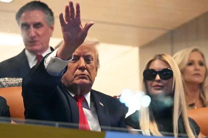 A public figure waves from a sports event booth; a woman with long hair and sunglasses sits next to him. Others are seated in the background