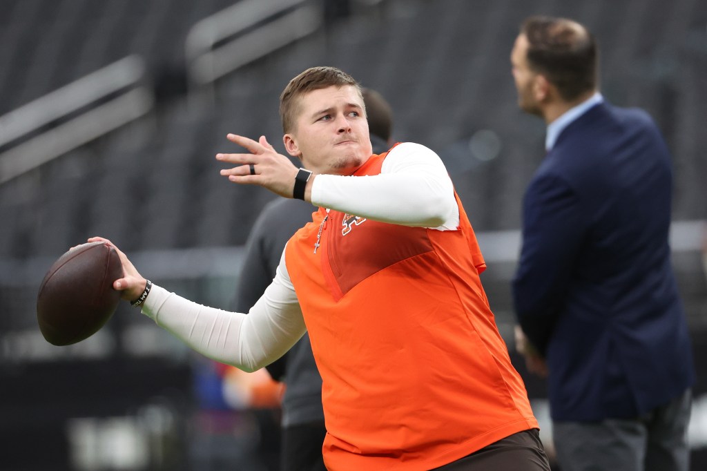 Cleveland Browns' Bailey Zappe warms up, wearing a white long-sleeved shirt under an orange vest and holding a football.