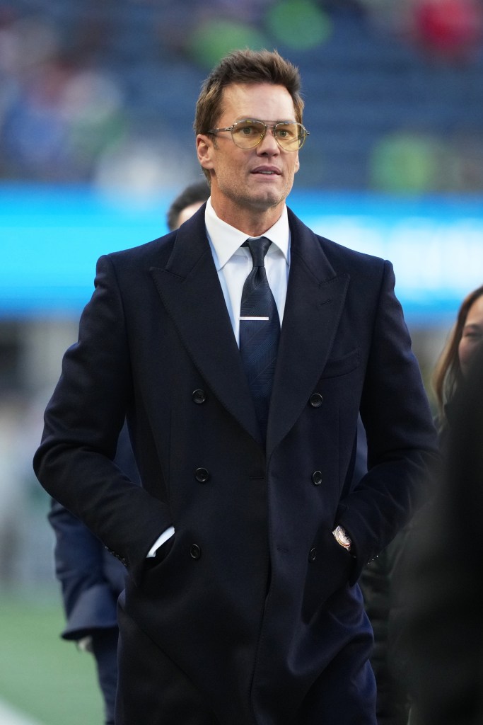 Tom Brady walking on a football field in a dark coat and yellow-tinted glasses.