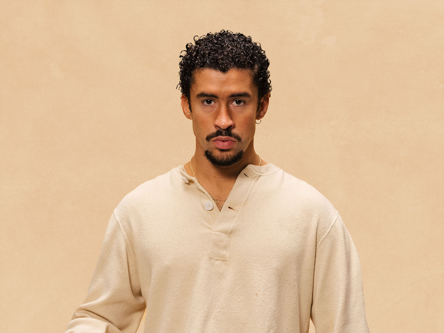 Bad Bunny, a male musical artist with dark curly hair and a goatee, wearing an off-white henley shirt.