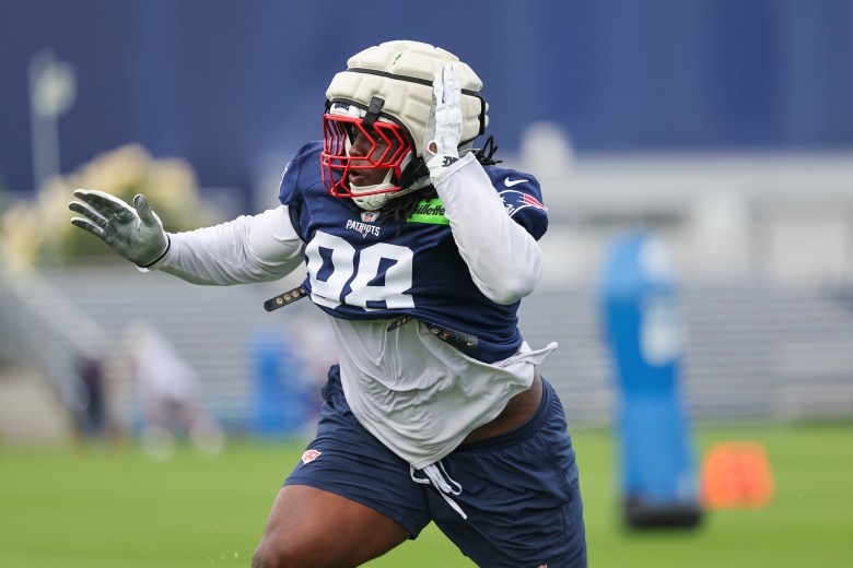 New England Patriots defensive lineman Jeremiah Pharms leans into a blocking sled during a practice drill, wearing a padded helmet and red facemask.