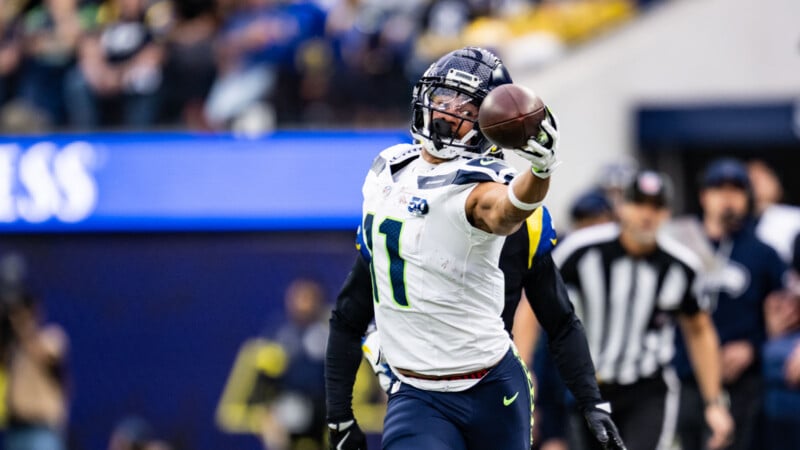 A football player in a white and navy uniform stretches his arm out while holding a football, celebrating during a game. Referees and other players are visible in the background.