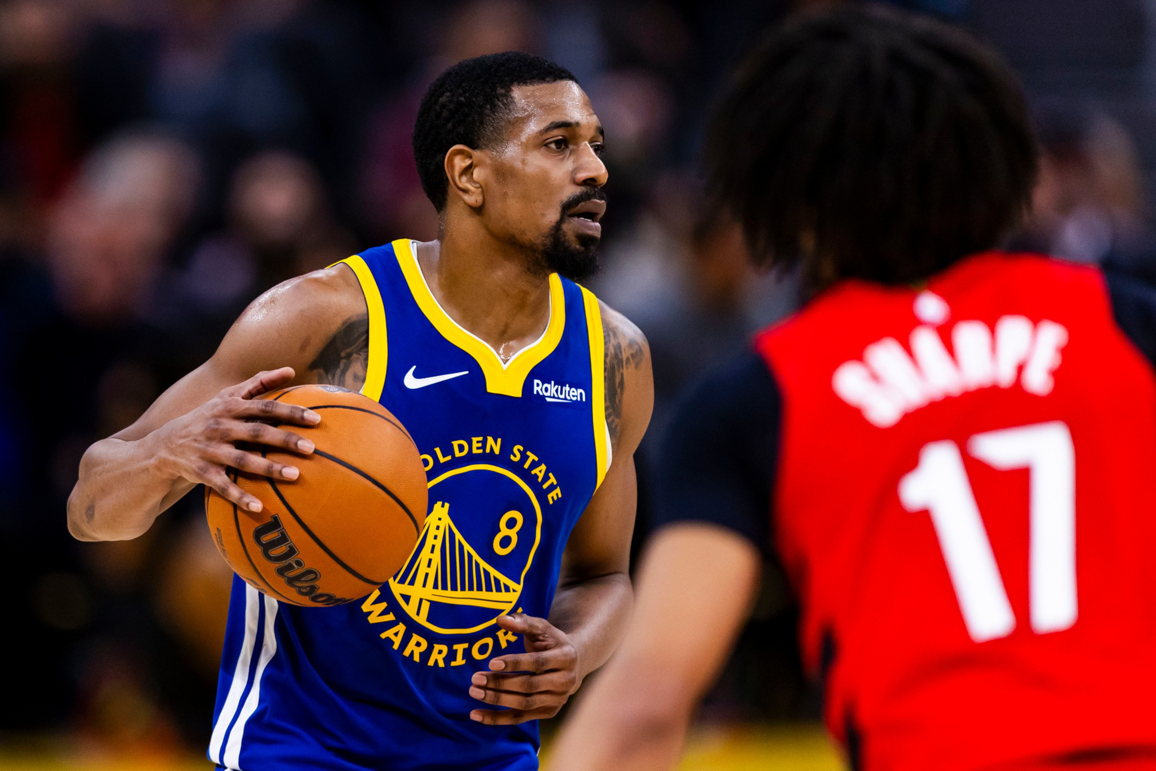 A Golden State Warriors player wearing a blue jersey dribbles a basketball while being guarded by an opposing player in a red jersey numbered 17.