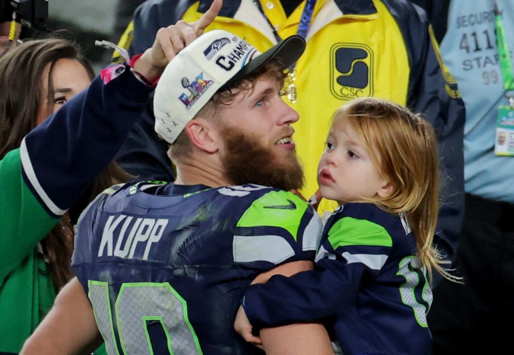 Seattle Seahawks' Cooper Kupp, in a championship hat and jersey, holding his daughter with his wife Anna Croskrey partially visible behind him.