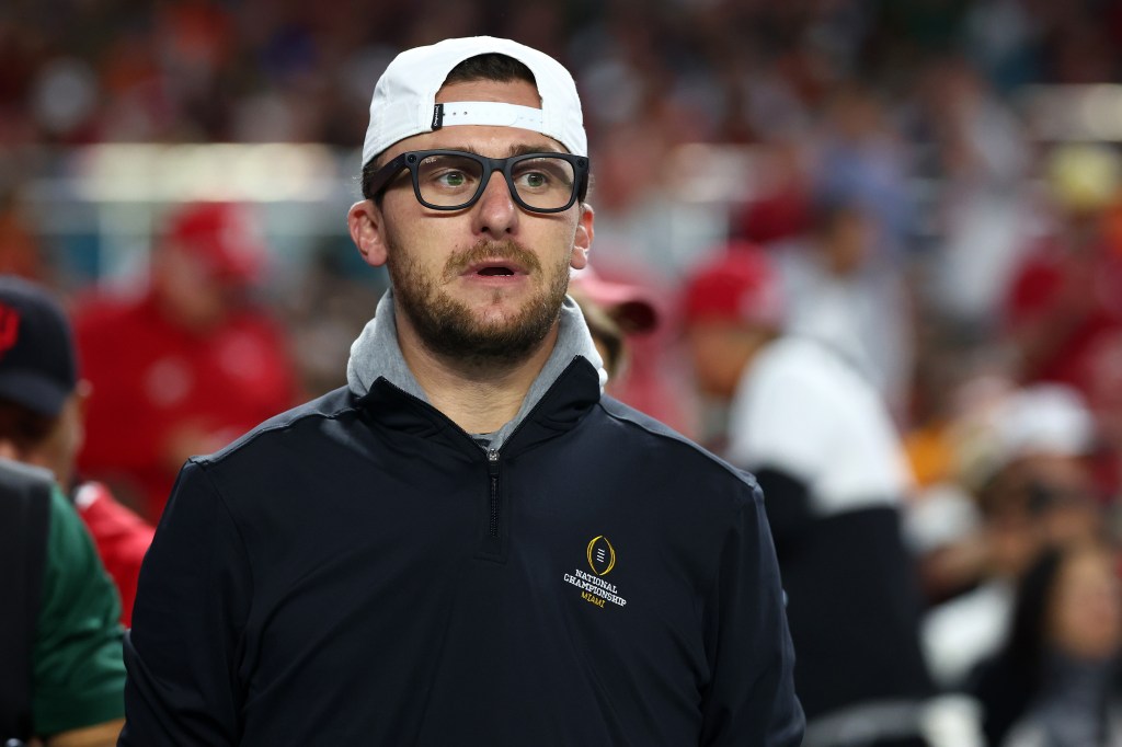 A man wearing a white cap backwards, glasses, and a black jacket with a "National Championship Miami" logo.