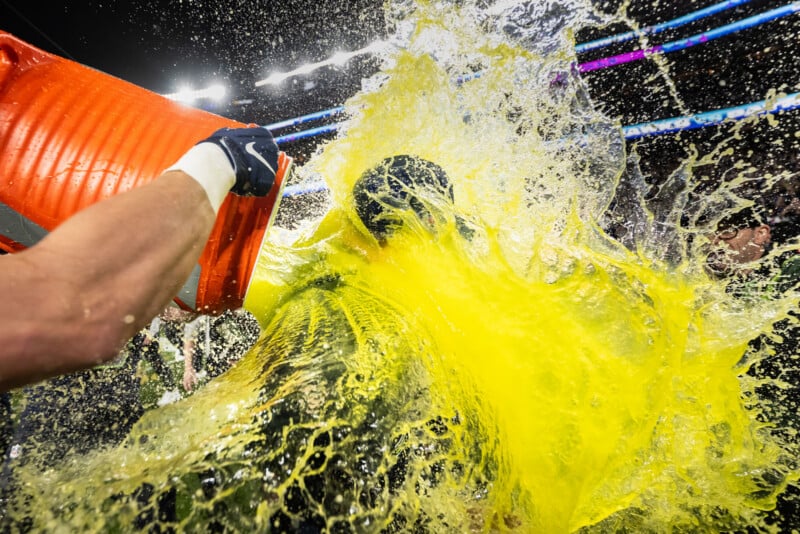 A person is being doused with a large splash of yellow sports drink from an orange cooler during a celebration on a brightly lit sports field.