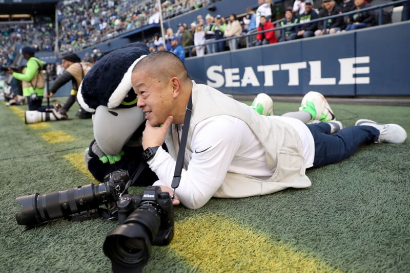 A man lies on a football field near the sideline, smiling with his hand on his cheek, next to a person in a large bird mascot costume. Two professional cameras rest on the ground in front of him. "SEATTLE" is visible on a wall behind them.