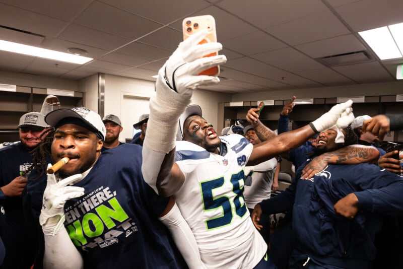 A group of Seattle Seahawks players celebrate in a locker room, smiling and cheering as one player in uniform takes a selfie with his phone. Some players hold cigars and wear victory T-shirts.