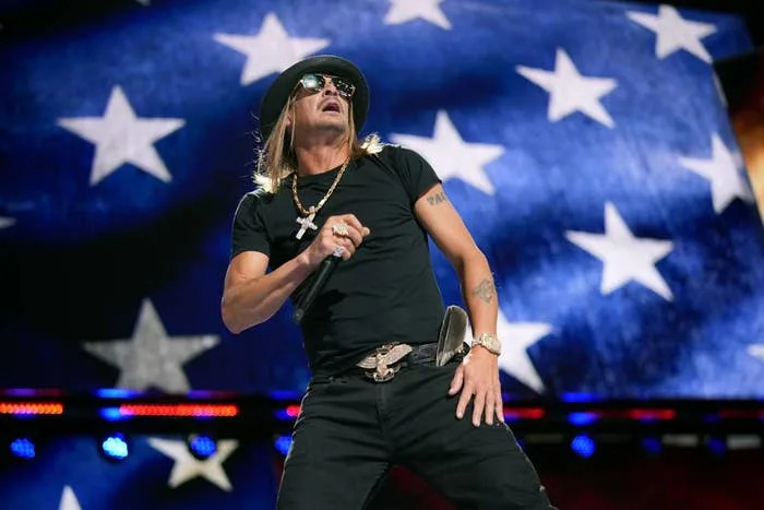 A performer in casual, edgy attire with a hat and sunglasses sings energetically onstage against a starry backdrop