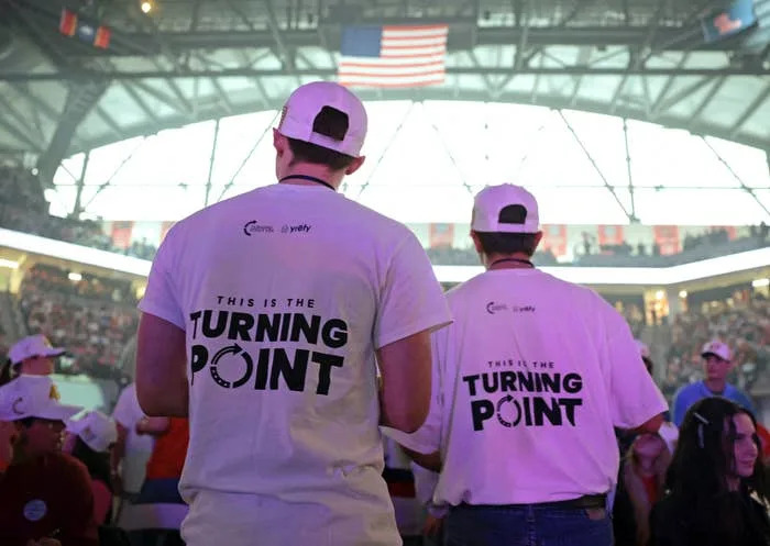 Two individuals wearing "This is the Turning Point" T-shirts at a crowded sports event under a large arena roof