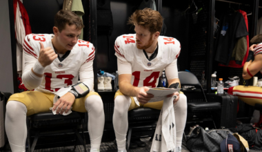 Two football players in San Francisco 49ers uniforms sit on chairs in a locker room, one talking while the other listens holding a towel.