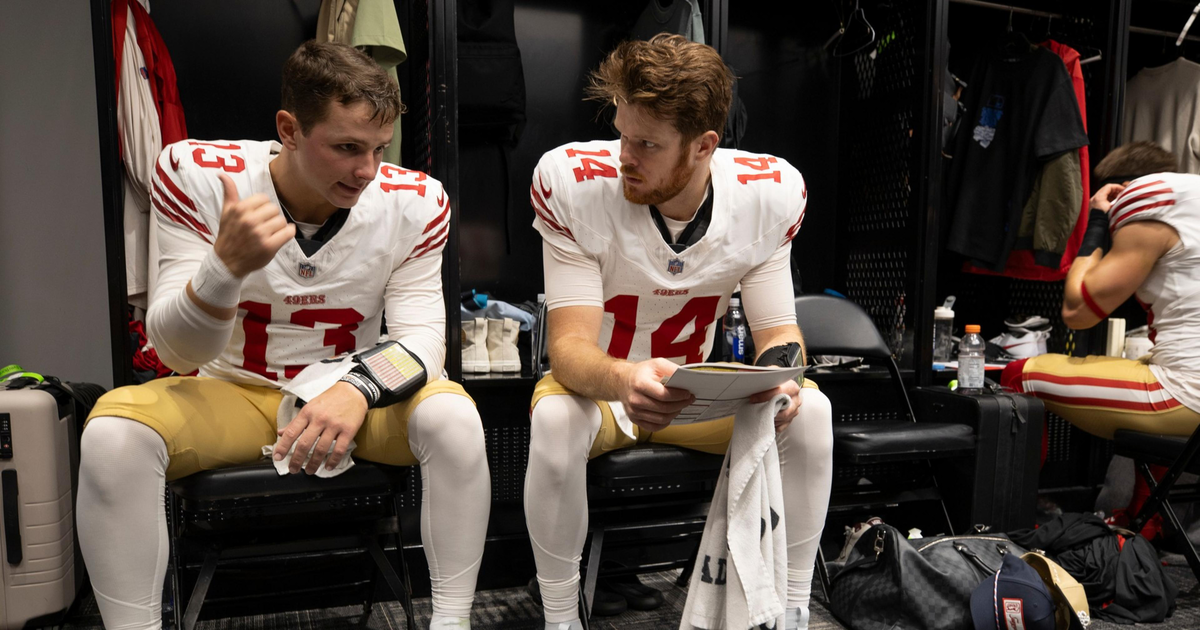 Two football players in San Francisco 49ers uniforms sit on chairs in a locker room, one talking while the other listens holding a towel.