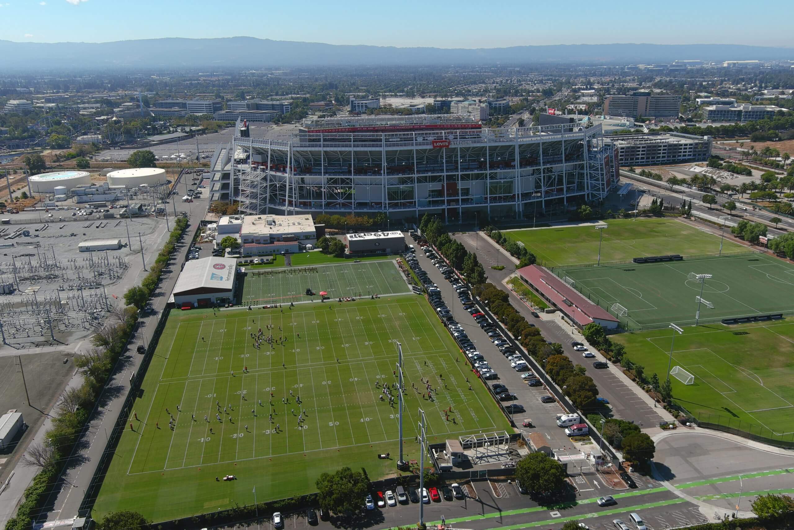 An aerial view of the 49ers' three practice fields (center left), with the weight room building at the left end of the third field. Levi's Stadium is beyond the fields, and the electrical substation is to the left across a road.