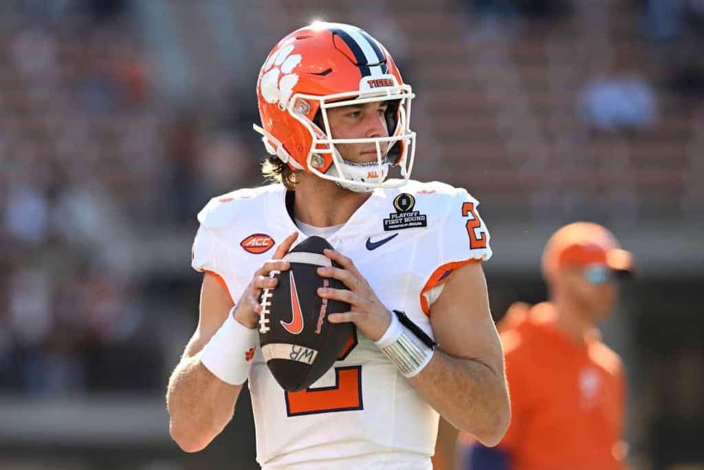 Clemson Tigers quarterback #2 scans the field before a snap, holding the football with both hands in a white road uniform.
