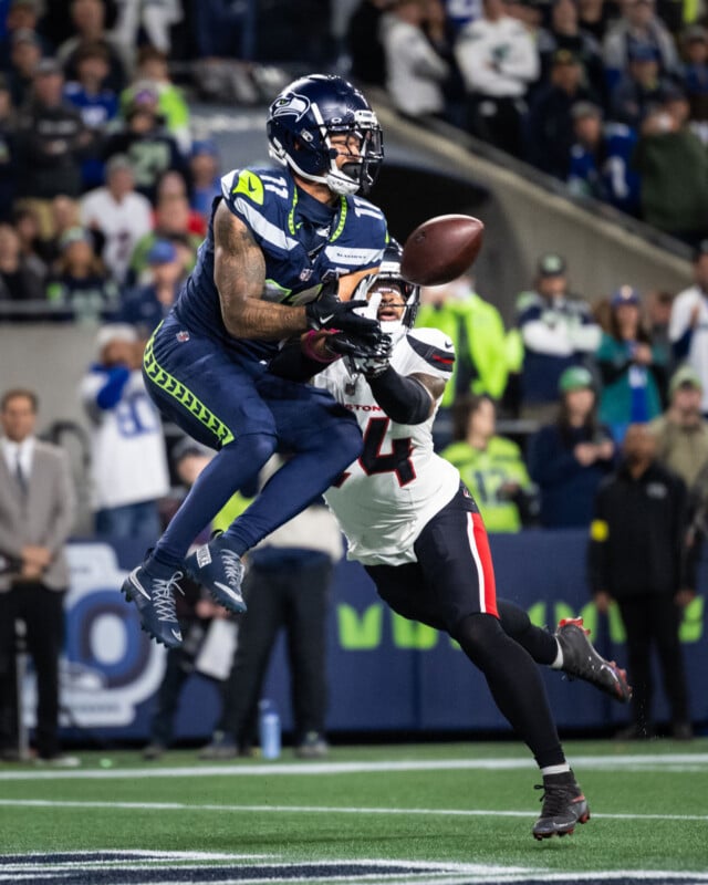 A Seattle Seahawks player jumps to catch a football while being closely defended by a player in a white and black uniform during an NFL game, with fans watching in the background.