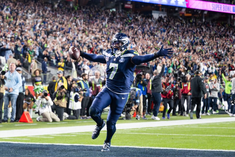 A Seattle Seahawks player in full uniform celebrates with arms wide after scoring a touchdown in front of a cheering crowd during a football game. Photographers and fans fill the stadium background.