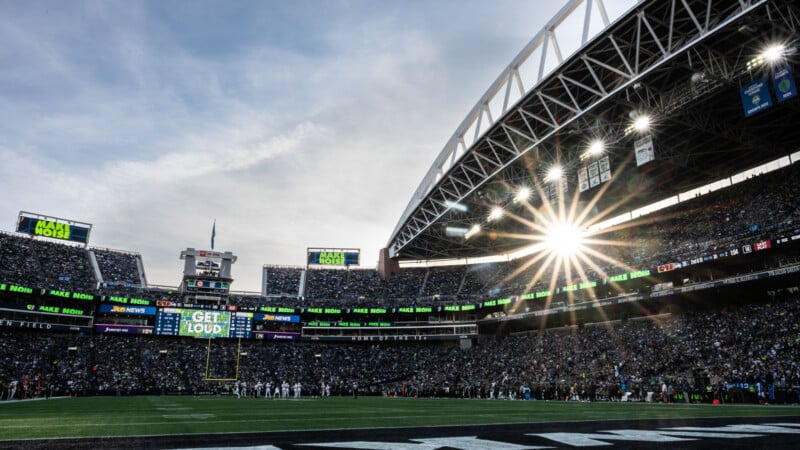 A packed football stadium under a partly cloudy sky, with sunlight streaming through the open roof. The scoreboard displays "GET LOUD" and fans fill the stands, cheering on the teams lined up on the field.