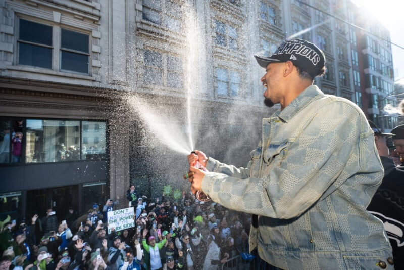 A man in a "Champions" hat sprays champagne over a cheering crowd during a street celebration, with people raising their arms and holding signs in front of tall city buildings.