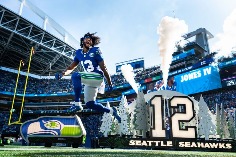 A Seattle Seahawks football player jumps energetically near a large “12” sign, with smoke and fans filling the stadium on a sunny day. The scene is vibrant, with team colors and excitement in the air.