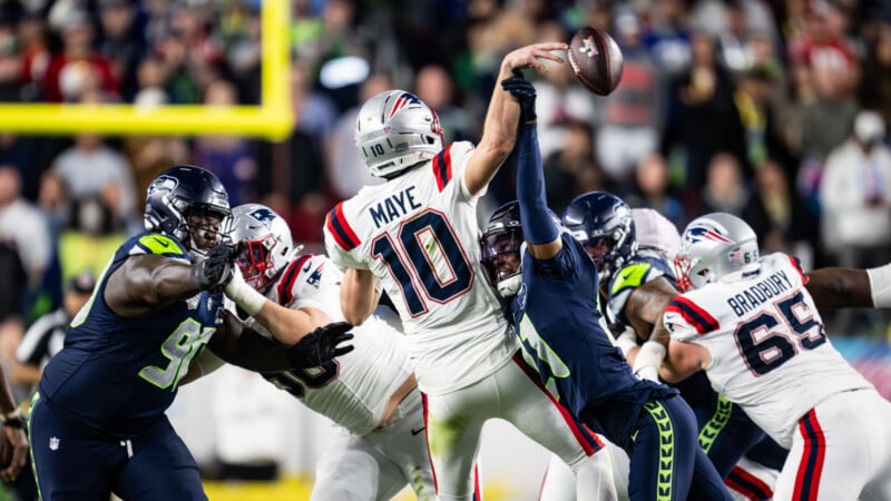 A football quarterback in a white uniform throws a pass while being pressured by defenders in dark blue uniforms during a game. Teammates and opponents are engaged in blocking and tackling around him on the field.