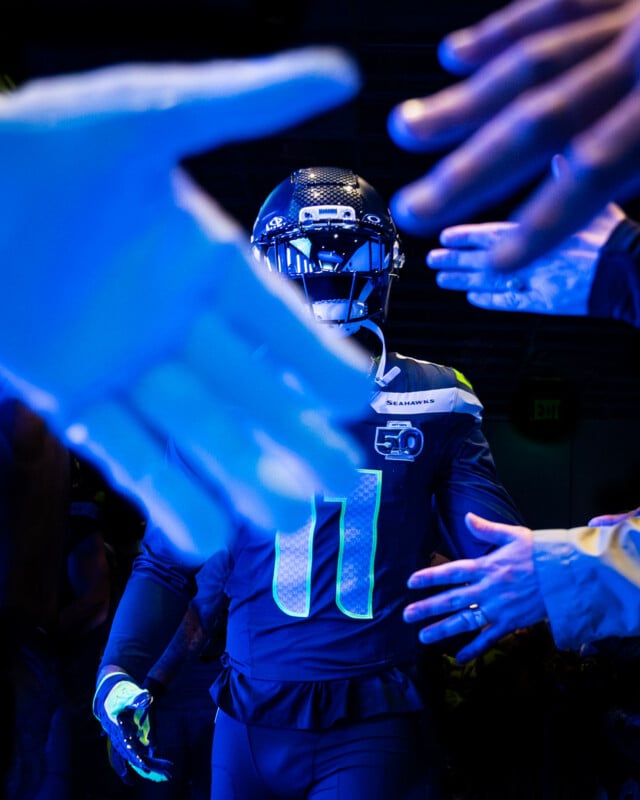 A football player in a dark uniform walks through a tunnel, reaching out to high-five fans whose hands are extended toward him under dramatic blue lighting.
