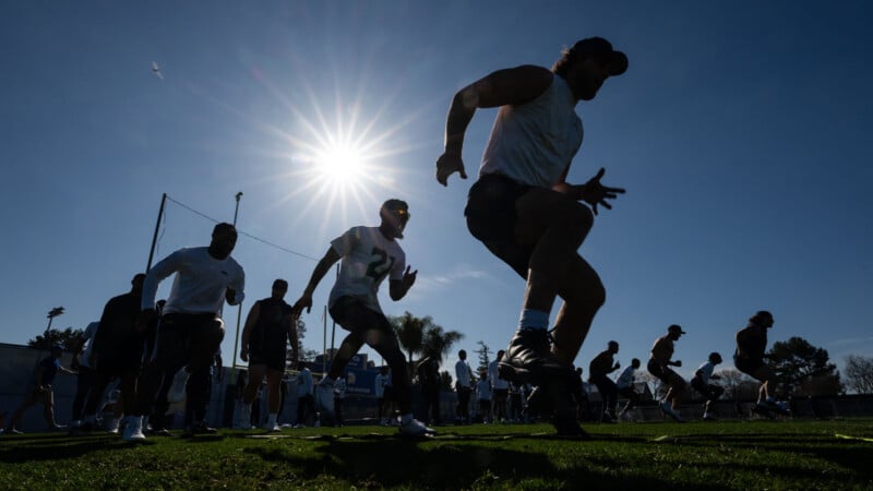 A group of athletes runs drills on a grassy field under a bright sun, casting long shadows as they train vigorously against a clear blue sky.