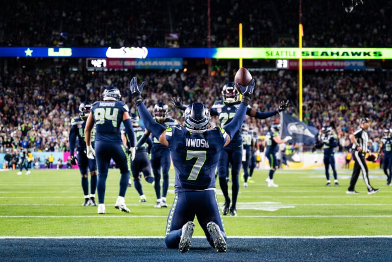 A Seattle Seahawks player, number 7, kneels on the field with arms raised holding a football, celebrating as teammates gather in the background during a night game with a cheering crowd.
