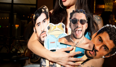 A person holds four cardboard cutouts on sticks: three male faces and one cartoon frog wearing a blue shirt.