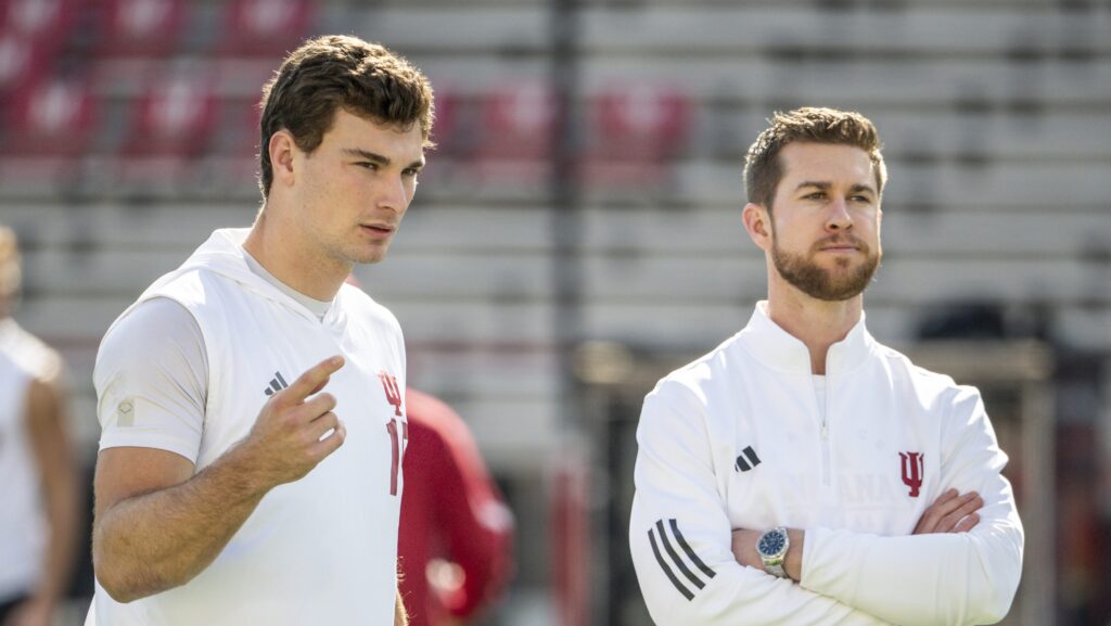 Two individuals in athletic attire stand on a football field, discussing strategies or observations. One is pointing while the other observes thoughtfully.