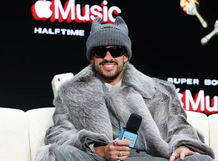 Person in a fur coat and beanie, wearing sunglasses, holding a microphone at a sports event press conference