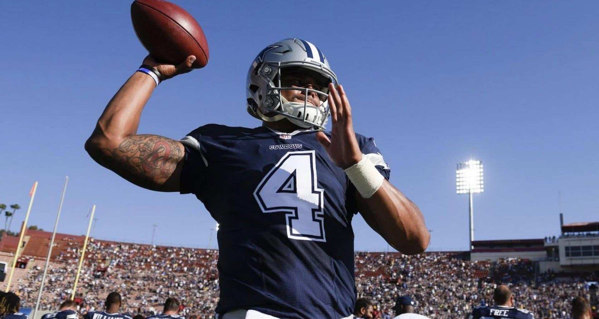 Aaron Rodgers in Dallas Cowboys football uniform, holding football on field during game, under stadium lights, showing athletic build and tattooed arm, focused on play.