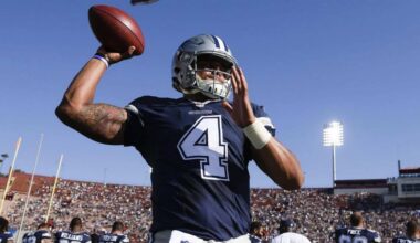 Aaron Rodgers in Dallas Cowboys football uniform, holding football on field during game, under stadium lights, showing athletic build and tattooed arm, focused on play.