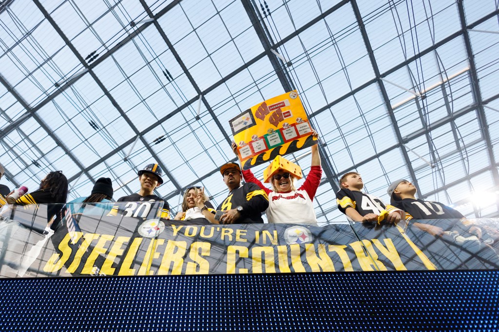 Fans of the Pittsburgh Steelers football team in the stands, with one fan holding a sign listing Steelers players and another wearing a cheesehead hat.