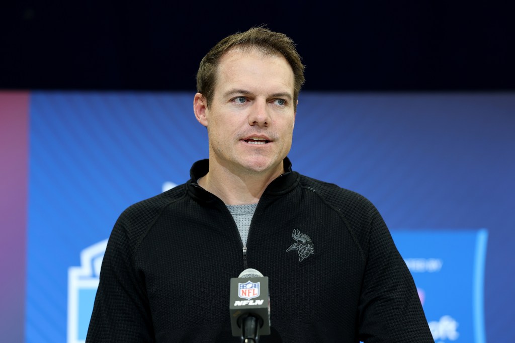 A man in a black and grey quarter-zip top speaking at a podium with an NFL logo microphone.