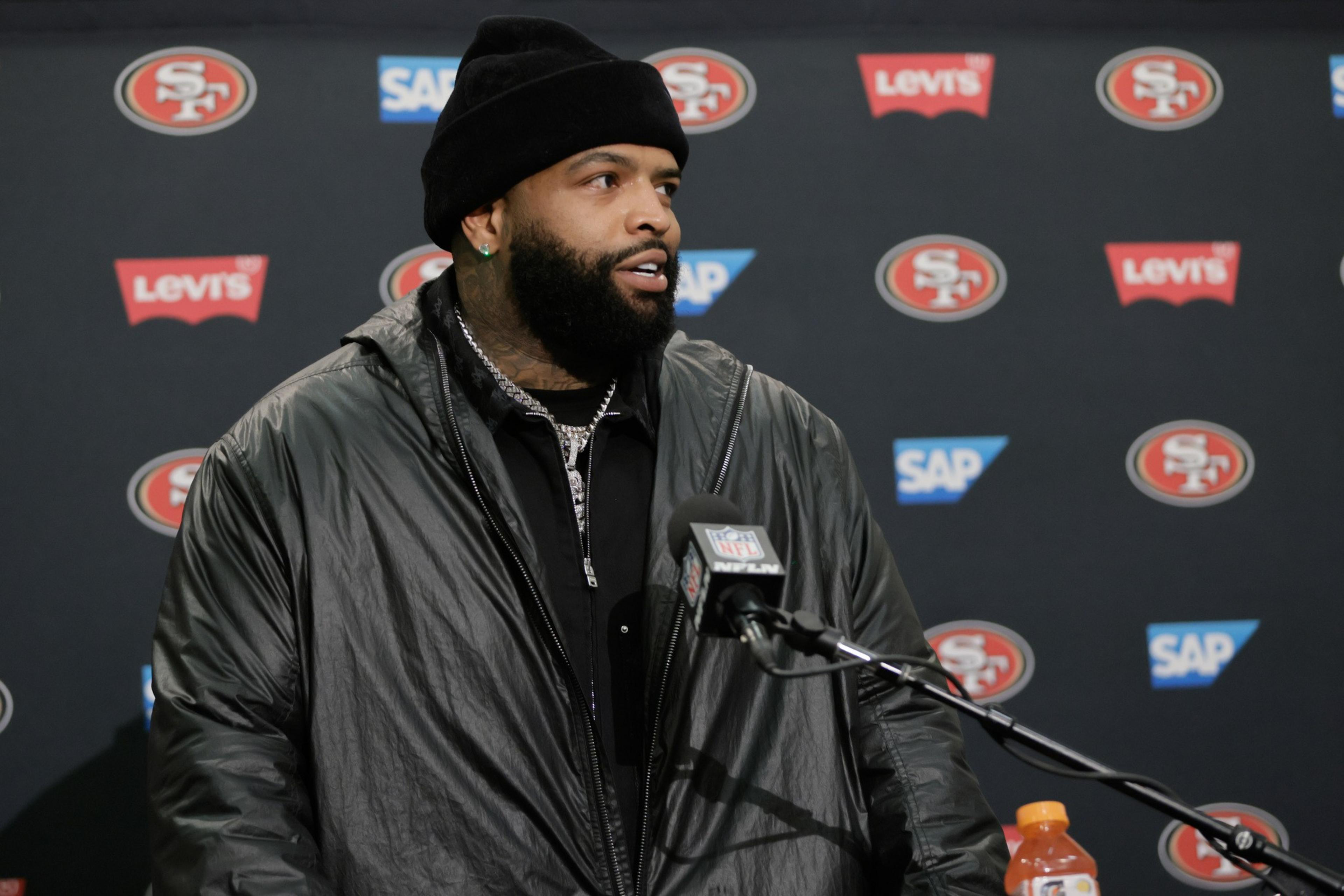 A man with a beard and black beanie speaks at a San Francisco 49ers press conference, wearing a large black jacket beside an NFL microphone.